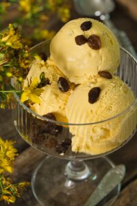 Three scoops of yellow ice cream topped with raisins in a glass bowl, outdoors.